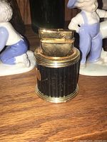 Front angled view of vintage Colibri table lighter showing gold-tone top and base with black ribbed cylindrical body, placed on wooden surface with decorative figurines in background