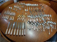 Wide assortment of multiple types of used stainless steel cutlery pieces arranged on a wooden table including forks, knives, large spoons, small spoons, and specialty utensils.