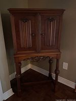 Front view of the wooden bar cabinet showing raised panel doors with carved details and round knobs, resting on a stand with carved legs and scrolling design.