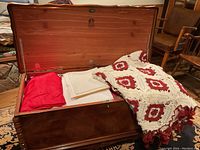 Open cedar chest showing inside with assorted linens including a red fabric, white folded linens, and a white/red crocheted blanket.