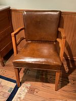 Front view of the brown leather armchair showing seat, backrest, wooden arms and legs, with wood panel wall background and hardwood floor