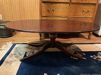 Full view of the oval coffee table showing the wooden top and pedestal base with curved legs on a floral and blue rug.