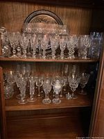 Mixed crystal glassware displayed on two wooden shelves, showing various stemmed and short glasses, with some decorative cut patterns.