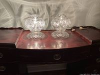 Pair of clear crystal table lamps on a dark wooden surface with their cords and switches visible, lit and showing reflective patterns on the wall.