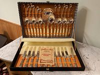 Full cutlery set in open case showing knives in bottom compartment and various forks, spoons, and serving utensils in upper lid compartment.
