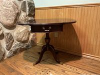 Wooden drop leaf table against stone and wooden wall, showing overall shape and style with legs and drawer visible.