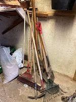 Photo showing multiple garden tools leaning against a wall, including rakes, shovels, and a broom with wooden handles and metal heads or bristles.