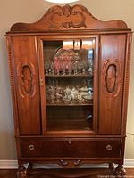 Front view of the wooden hutch showing glass center door with shelves filled with glassware, wooden side doors with carved handles, and bottom drawer with round pulls.