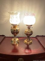 Two vintage brass oil lamps side-by-side on wooden surface, both lit showing the glass shades, one with clear etched scalloped glass, the other with white and pink ruffled glass.