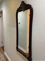 Side angle view of the tall rectangular mirror showing ornate carved dark wooden frame with a crest design at the top and slight frame wear.