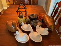 Overview of all decorative items arranged on a wooden table showing four milk glass hen dishes, wooden duck decoy, metal trays, ceramic lantern, and glass bowls.