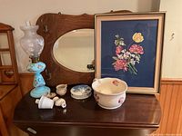 Wide view of all lot contents on dark wood table; includes oil lamp, floral artwork, ceramic items and mirror visible in background.