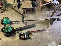 Three lawn and garden power tools including string trimmer, leaf blower, and hedge trimmer on a concrete floor surrounded by workshop items.