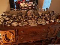 Wide angle view of all silverplate tableware items arranged on a wooden sideboard, showing the variety and number of pitchers, trays, bowls, and other pieces.
