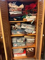 Open wooden storage cabinet showing stacked assorted folded fabrics on top shelf and multiple plastic containers filled with sewing supplies on lower shelves.