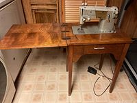 Singer sewing machine mounted in wood cabinet with folding top cover open to left showing sewing machine and drawer below