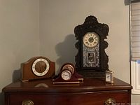 Front view of all four clocks on a wooden surface against a pale wall showing the tall ornate wooden mantle clock with patterned etched glass, two wooden mantle clocks, and a small cylindrical clock.