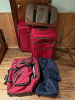Photo showing five travel bags: brown soft briefcase on top, two red upright suitcases, a large red duffel bag, and a large navy duffel bag on floor