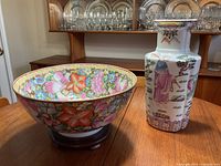 Full view of both porcelain items, bowl and vase, on wooden table with cabinet and glassware in background.