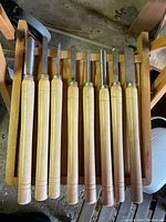Front view of 9 woodworking chisels with wooden handles resting on a wooden rack, showing the handles and blade bases.