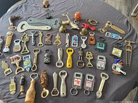 Wide-angle photo showing a large assortment of metal and plastic bottle openers with various shapes and sizes arranged on a cloth surface.