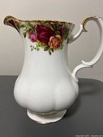 Side profile of vintage milk jug showing floral rose pattern and gold trim on white bone china with scalloped body shape and handle.