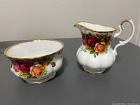 Photo showing the creamer and sugar bowl side by side on a flat surface, highlighting the floral pattern and gold trim.