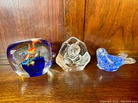 Photo of three glass paperweights on wooden surface showing colorful fish in vase, frosted bear, and blue bird designs.