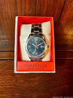 Front view of the Caravelle men's dress watch inside original orange Caravelle New York box showing black dial with Roman and stick hour markers, date window, and stainless steel bracelet.