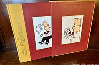 Front and spine view of the 'Dilbert 2.0 - 20 Years of Dilbert' hardcover book with slipcase showing cover art of Dilbert holding Dogbert and gold embossed 'S. Adams' on spine.