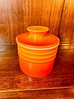 Front view of the orange stoneware butter bell crock with lid on wooden surface showing glossy finish and ring design.