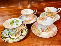 Four Royal Albert fine bone china tea cups with matching saucers showcasing varying floral and scenic patterns, all arranged on a wooden surface.