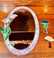 Overview of the oval mirror with iridescent pink glass segments framing and three pink roses with green leaves decoration. Shows additional loose stained glass pieces: a blue mushroom, a green flower, and two mushrooms in brown and tan.