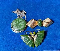 Three pieces of sterling silver and jade jewelry displayed on blue fabric: Asian pendant with etched jade longevity symbol on chain, butterfly brooch with jade wings, and bar brooch with jade cabochon.