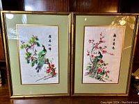 Pair of framed vintage Chinese silk embroidery artworks of peacocks on branches under glass, with bamboo-style gold frames on wooden surface.