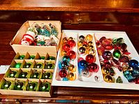 Large assortment of vintage holiday decorations and Christmas bulbs displayed on a table including boxed green bulbs, assorted colored bulbs in a tray, and a box with diverse decorative pieces.