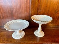 Two porcelain compotes displayed on a wooden surface; left is Wedgwood with embossed floral design, right is Lenox with sunflower petals and gold trim.