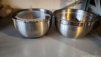 Two large stainless steel mixing bowls nested together with a large clear measuring cup inside one bowl, placed on a shelf.