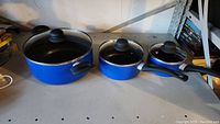 Three blue nonstick pots of large, medium, and small sizes with black handles and glass lids with black knobs, shown closed.