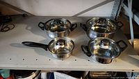 Four stainless steel pots with black handles and glass lids displayed together on a shelf.