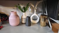 Five decorative vases on a shelf including pink, cream, dark, white, and gold colored vases