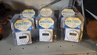 Photo showing six 7-day dual outlet digital timers in original packaging, displayed on a shelf.