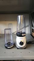 Starfrit electric blender base with one long blending container attached and one shorter container beside it on shelf.