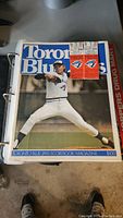 White binder with a Toronto Blue Jays pitcher on the cover and two game tickets inside the front plastic cover.