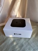 Closed Apple TV HD box in white with image of the Apple TV device on top and clear labeling on the front side.