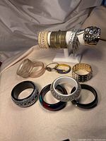 Photo shows multiple vintage 90s bangles arranged on a beige surface, including gold-tone textured bangles on a white display fixture, a studded silver-tone spiked bangle, translucent pink plastic bangles, and multiple colored and black bangles in front.