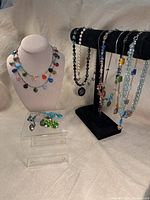 Photo showing a display of multiple colorful necklaces on a black velvet stand and a white bust, along with earrings on a clear stand below.