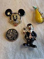 Close-up image showing three brooches: Mickey Mouse head in black and gold with rhinestones, a round monogram button brooch, and a clown figure in black suit with red tie and cane.