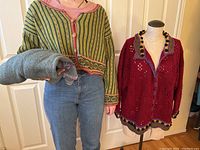 Model wearing green striped cropped knitted sweater with floral edges and buttons holding grey knitted sweater, red open knit cardigan on mannequin nearby.