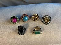 Group of six vintage cocktail rings arranged on light grey fabric, showing all rings with various colorful stones and rhinestone details